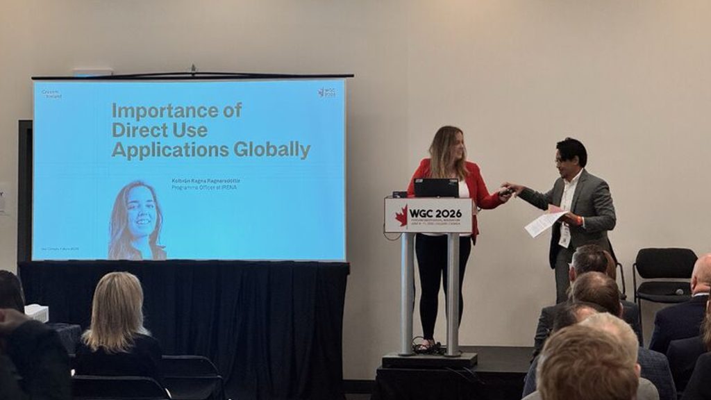 Presenter at a podium hands over a token to a colleague beside a slide reading 'Importance of Direct Use Applications Globally' with a projected screen and audience watching