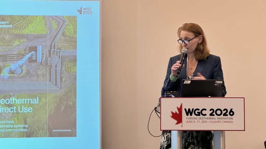 Female presenter in a navy blazer speaks into a microphone at a podium with a WGC 2026 banner/poster beside her, during a geothermal conference