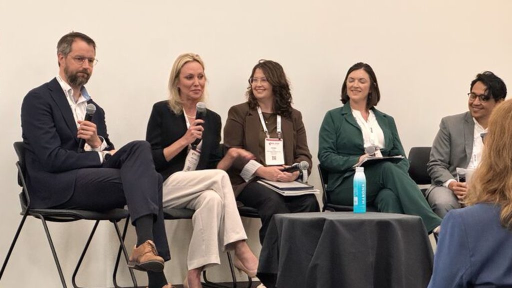 Panel of five professionals seated in a row, each holding a microphone, participating in a conference discussion in a neutral room.