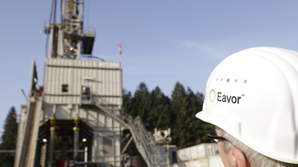 Eavor hard hat | ThinkGeoEnergy - Geothermal News & Insights Person wearing a white Eavor hard hat in foreground surveying a large industrial drilling rig at a construction site.