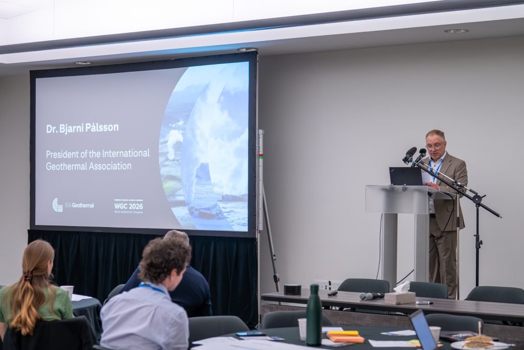 Gas management Bjarni | ThinkGeoEnergy - Geothermal News & Insights Man in a suit giving a talk at a conference podium with a large screen displaying the speaker's name, Dr. Bjarni Pálsson, President of the International Geothermal Association, in the background.