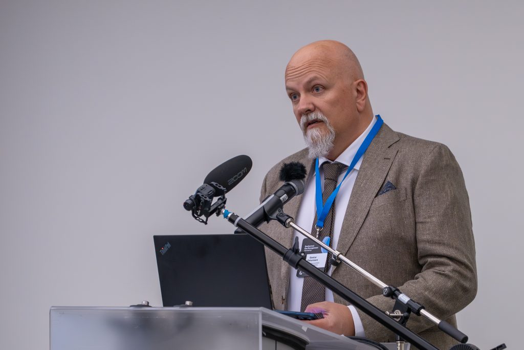 Gas management Gestur | ThinkGeoEnergy - Geothermal News & Insights Man in a brown suit speaking at a podium with multiple microphones during a formal event, wearing a blue lanyard and tie