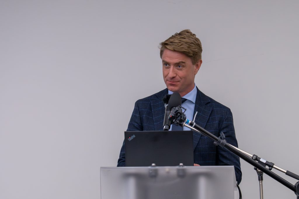 Gas management Jonas | ThinkGeoEnergy - Geothermal News & Insights Man in a blue check suit speaking at a podium with a laptop and multiple microphones in front of him.