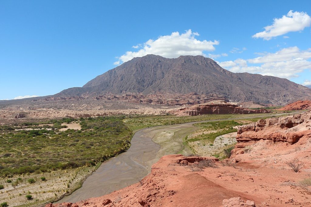 Desert valley with a muddy river winding toward a rugged mountain under a blue sky with white clouds.