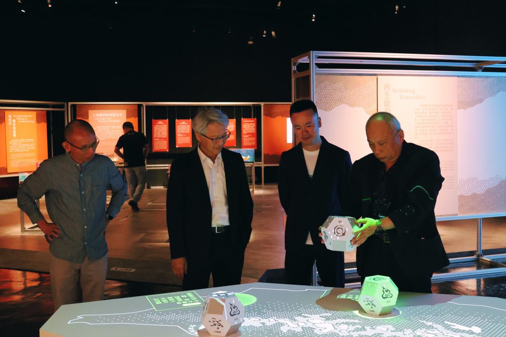 Energy planning | ThinkGeoEnergy - Geothermal News & Insights Four men in business attire stand around a lit exhibition table examining geometric dice on a map-like display, with orange information boards in the background.