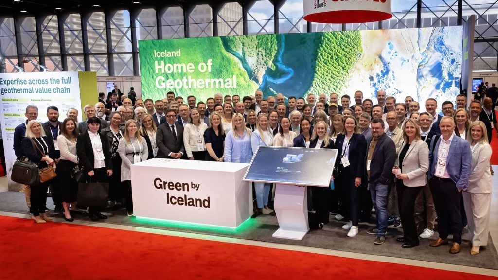 Green by Iceland delegation | ThinkGeoEnergy - Geothermal News & Insights Large group of professionals posing at an Iceland geothermal exhibit with banners reading 'Home of Geothermal'.