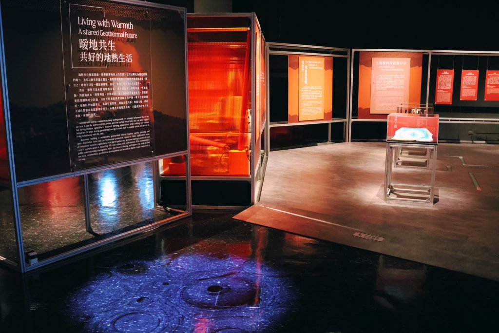 Living with warmth | ThinkGeoEnergy - Geothermal News & Insights Exhibition hall with black walls and orange-lit display panels about geothermal energy, including a large 'Living with Warmth' panel on the left and a glass display case in the foreground.