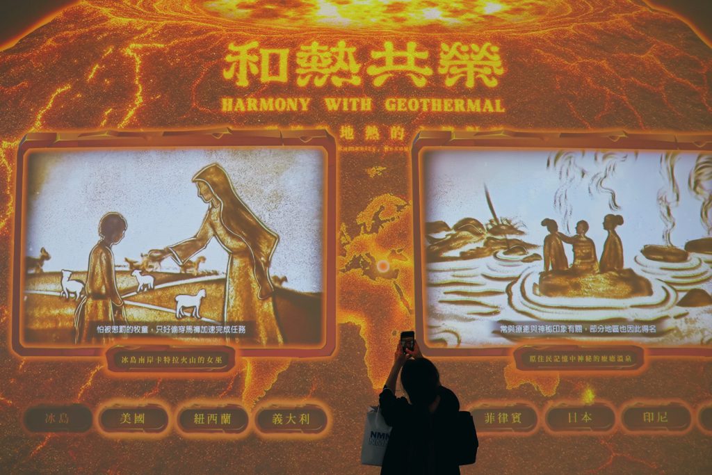 Memories of warmth | ThinkGeoEnergy - Geothermal News & Insights A person taking a photo of a bright orange exhibit wall with three glowing screens showing scenes and Chinese text about harmony with geothermal energy.