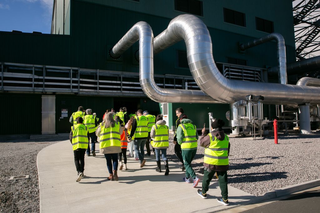 NZGW Contact Energy | ThinkGeoEnergy - Geothermal News & Insights Group of people in high-visibility jackets walk toward an industrial building with large silver ductwork.