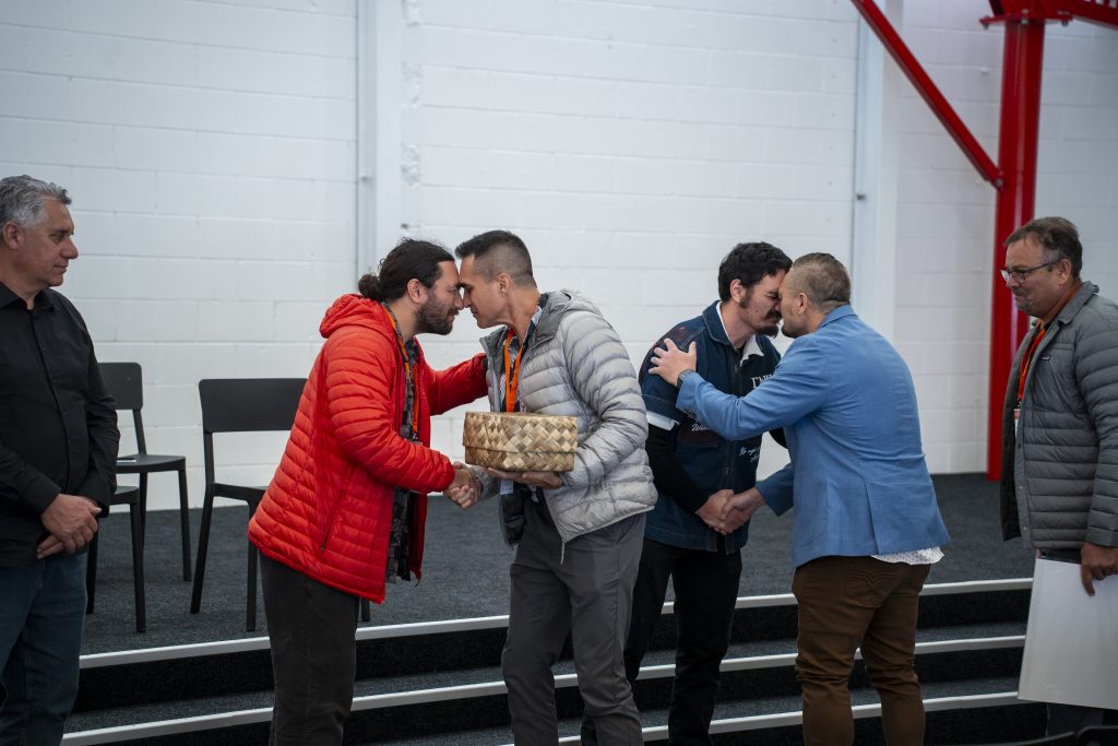 NZGW Maori | ThinkGeoEnergy - Geothermal News & Insights Two men greet with foreheads touching and a handshake, one in a red jacket and the other in a silver jacket, holding a woven basket between them, indoors.
