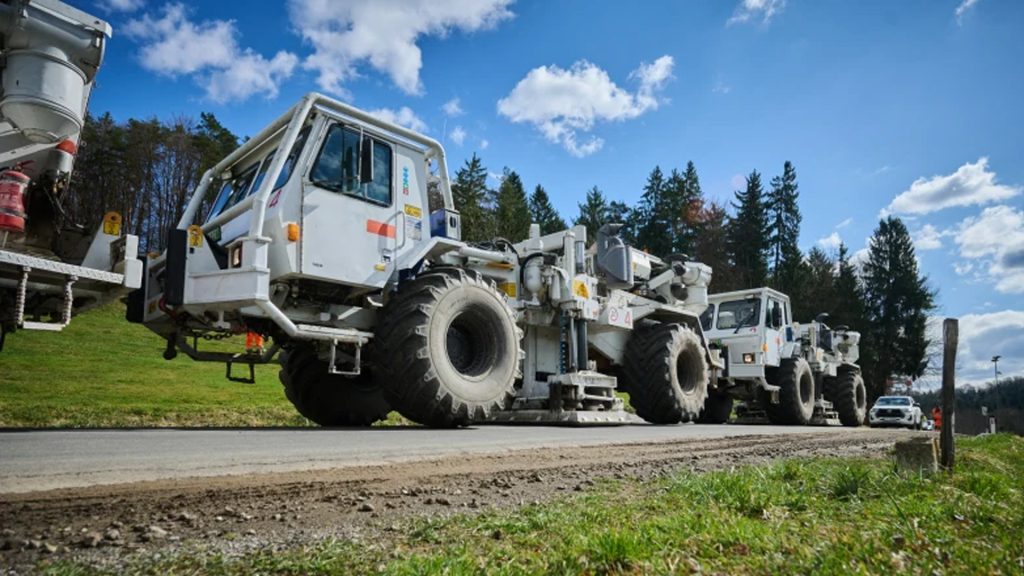 OMV Steiermarkt 1 | ThinkGeoEnergy - Geothermal News & Insights Line of large white industrial trucks with oversized tires parked along a road, under a blue sky with scattered clouds and trees in the background.