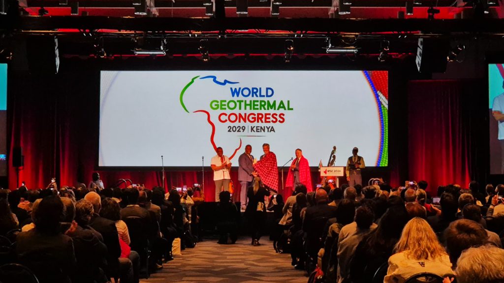 WGC Kenya handover | ThinkGeoEnergy - Geothermal News & Insights Onstage at the World Geothermal Congress 2029 Kenya, speakers stand in front of a large screen displaying the event logo as the audience watches.
