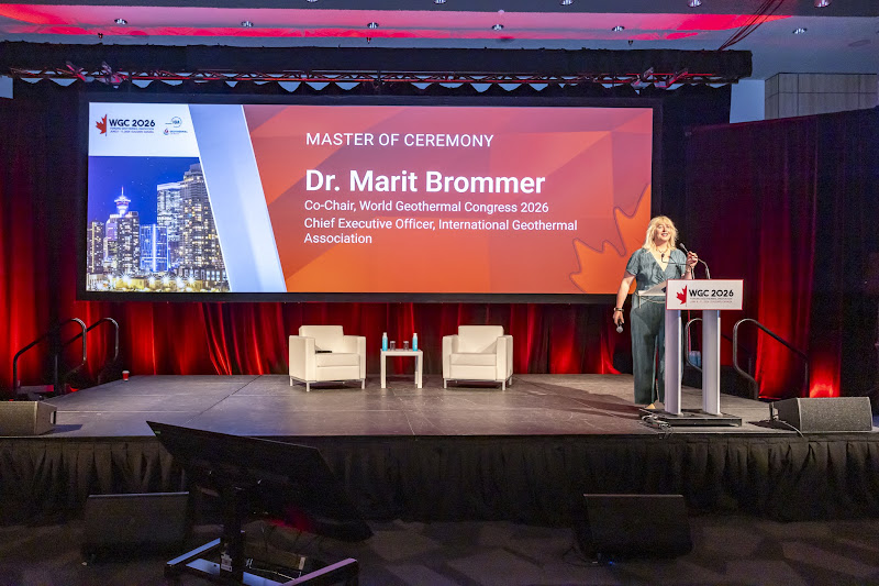 WGC Marit | ThinkGeoEnergy - Geothermal News & Insights Speaker at a podium on a stage with a large screen announcing 'Master of Ceremony' Dr. Marit Brommer at WGC 2026, red background lighting.