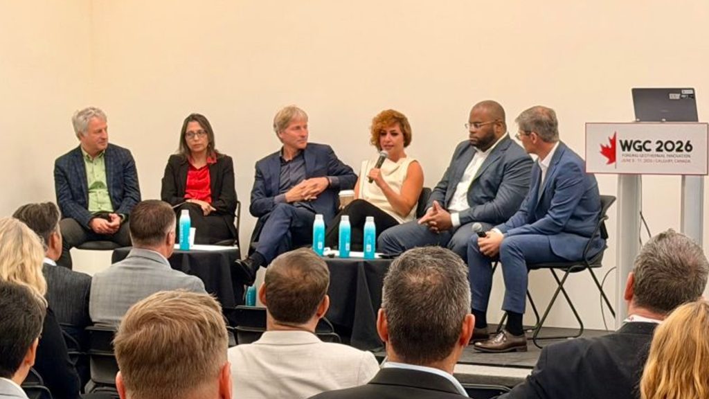 WGC panel Baker Hughes | ThinkGeoEnergy - Geothermal News & Insights Panel of six professionals seated on stage during a conference, with water bottles on small tables and a WGC 2026 podium nearby.