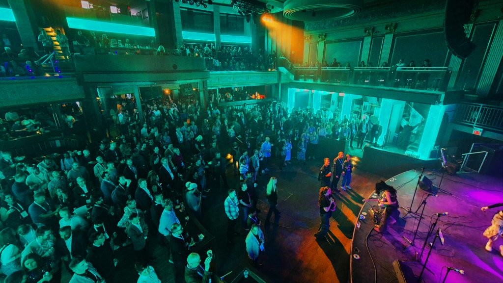 WGC_BOTB_023 edit | ThinkGeoEnergy - Geothermal News & Insights Large crowd fills a two-level club as a band performs on a brightly lit stage to the right, with teal and purple lighting.