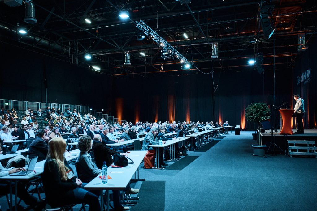 messe-offenburg_geotherm-2026_125249 | ThinkGeoEnergy - Geothermal News & Insights A conference hall with a speaker at a podium on stage, addressing a large seated audience.