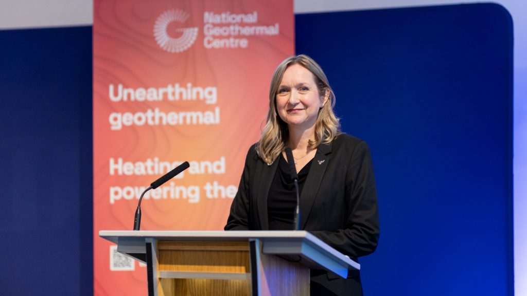 CAdams Headshot | ThinkGeoEnergy - Geothermal News & Insights Professional woman in a black blazer delivering a talk at a geothermal conference, standing at a podium with a red banner behind her