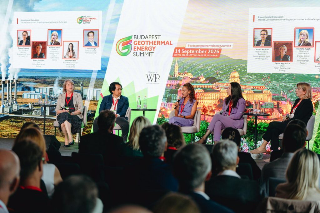Budapest_Geothermal_Summit_100_web | ThinkGeoEnergy - Geothermal News & Insights Panel discussion at a geothermal energy summit with five speakers seated on stage and an audience watching, backdrop shows 'Budapest Geothermal Energy Summit' and cityscape imagery.