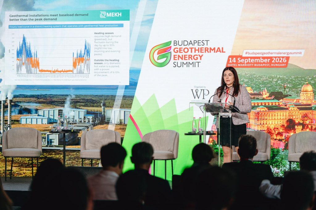 Budapest_Geothermal_Summit_124_web | ThinkGeoEnergy - Geothermal News & Insights Speaker at a podium presenting at the Budapest Geothermal Energy Summit, with a chart slide visible on stage left and an audience in foreground.