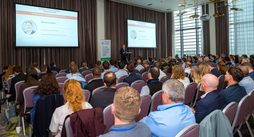 Darragh Dublin | ThinkGeoEnergy - Geothermal News & Insights Conference room full of attendees listening to a speaker at a lectern with two large presentation screens on either side of the stage.