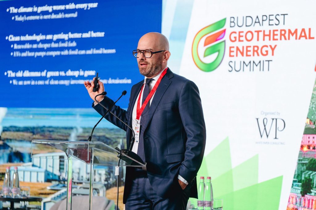 Istvan Bart | ThinkGeoEnergy - Geothermal News & Insights Speaker in a dark suit and glasses delivers a talk at a podium during the Budapest Geothermal Energy Summit, with a blue slide and summit branding in the background.