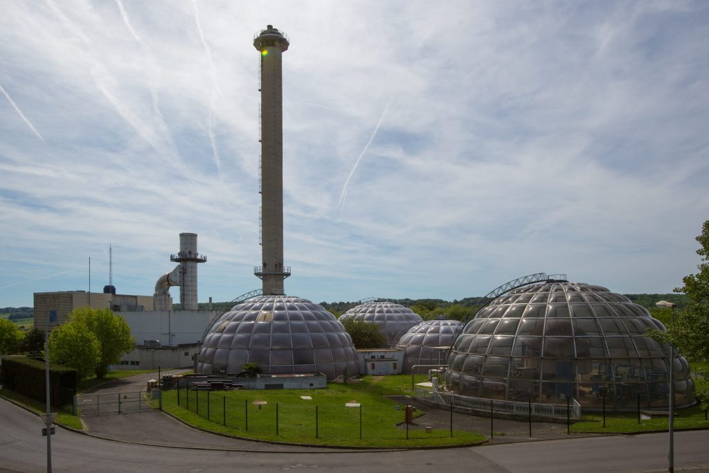 Meaux energy center | ThinkGeoEnergy - Geothermal News & Insights Industrial complex with a tall smokestack and dome-shaped glass buildings under a blue sky.