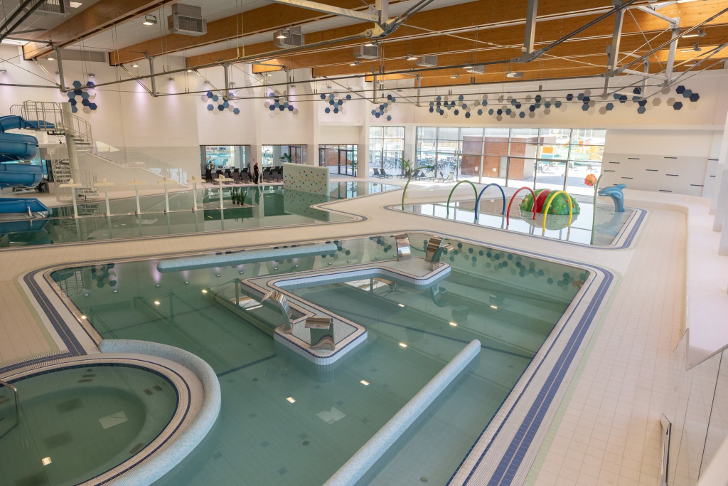 Wellover opening 2 | ThinkGeoEnergy - Geothermal News & Insights Indoor water park area with blue spiral slides on the left, multiple pools, and a glass wall showroom at the back.